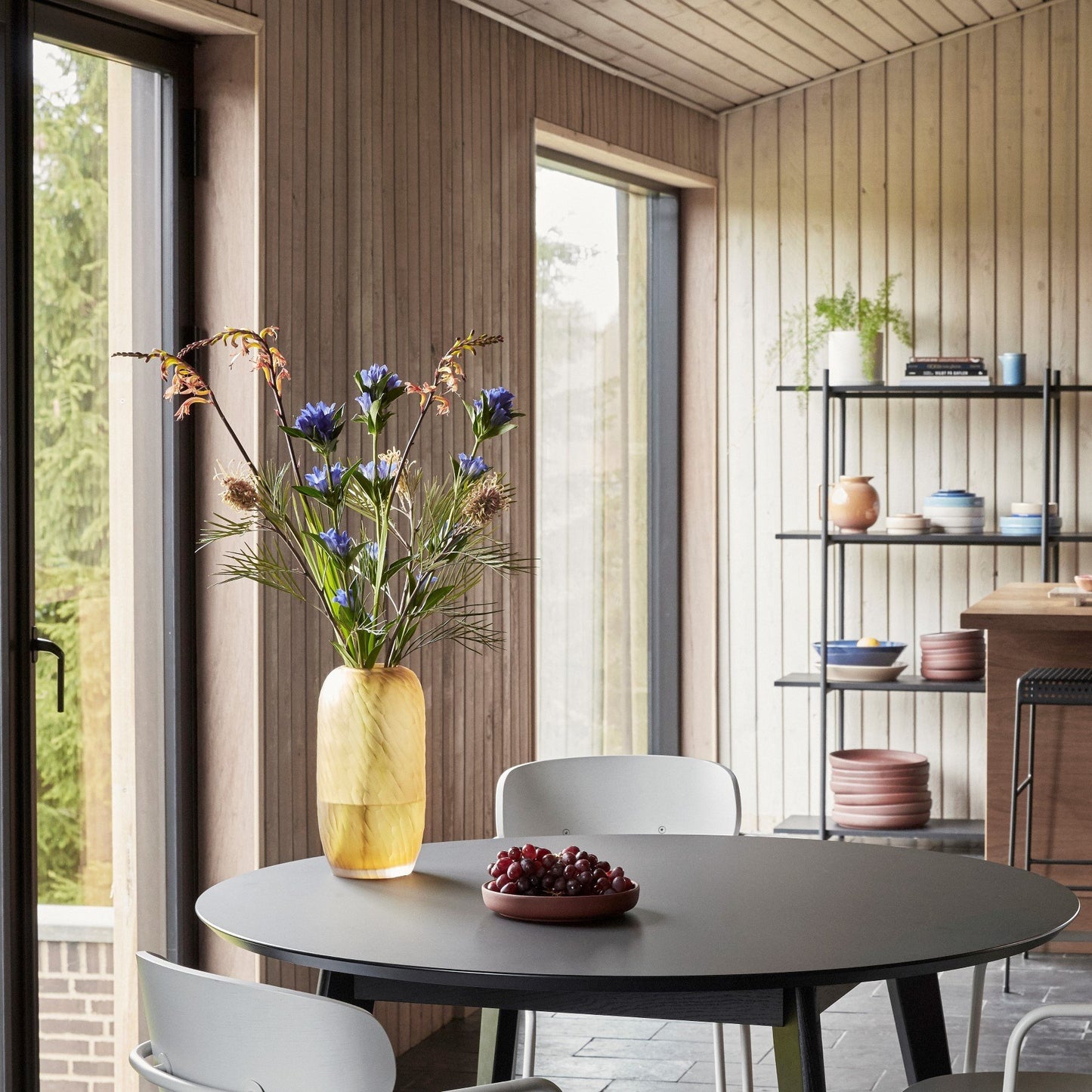 Hubsch Interior Scandi style tall dune textured vase in amber gold glass on a black wood dining table with flower arrangement