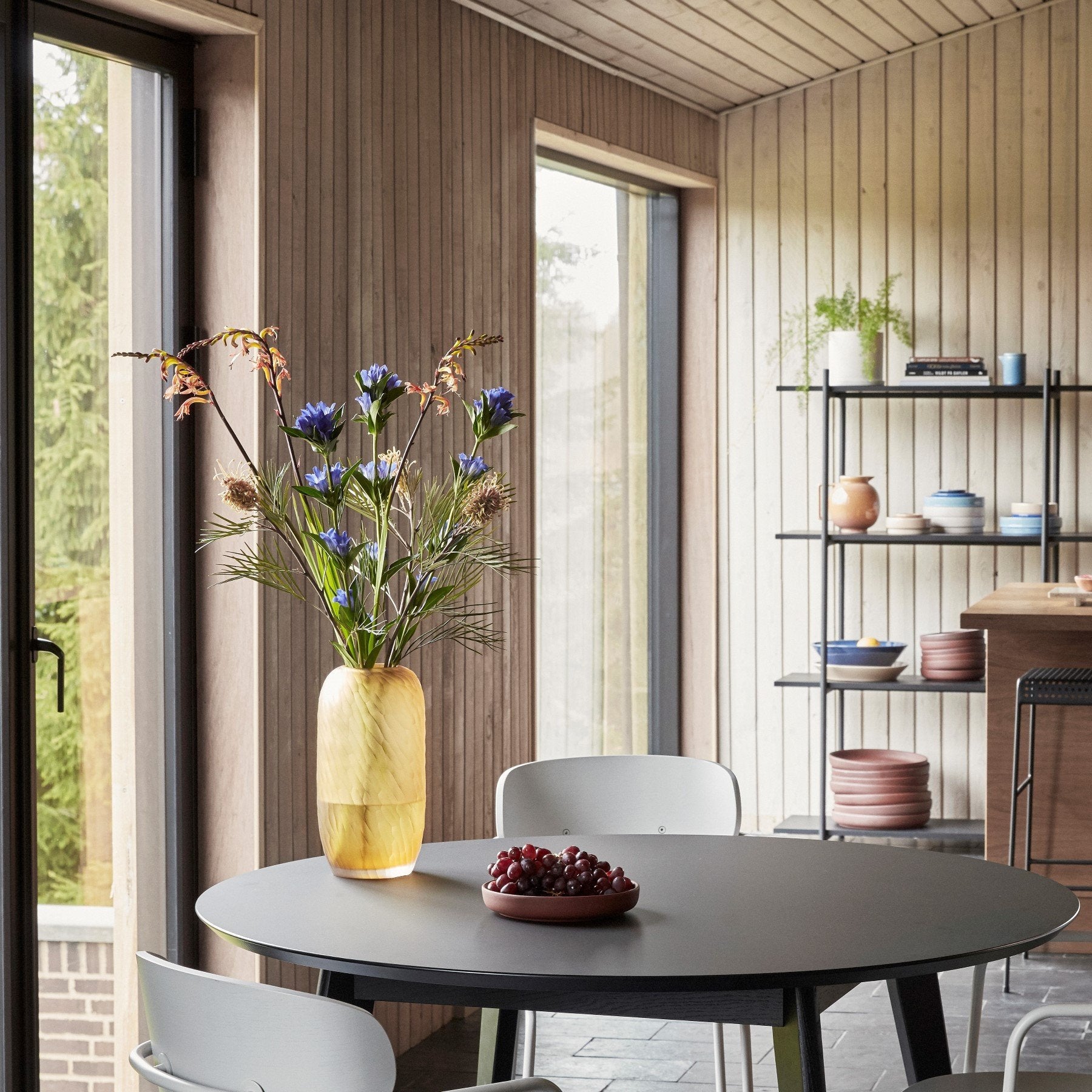 Hubsch Interior Scandi style tall dune textured vase in amber gold glass on a black wood dining table with flower arrangement